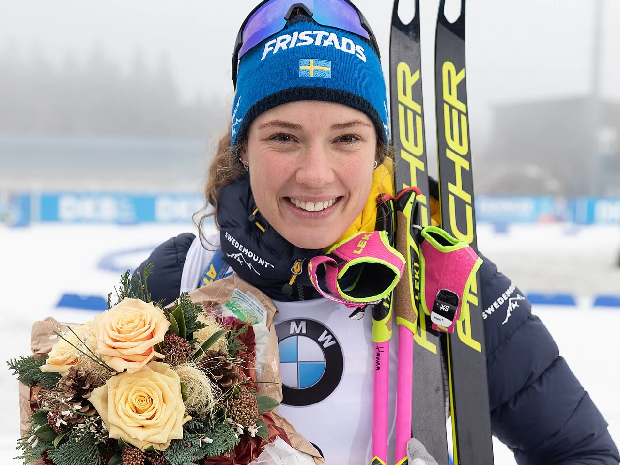 Hanna Öberg ler i blå mössa med skidor och en blombukett efter en världscuptävling i Oberhof
